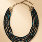 A multi-strand statement necklace featuring faceted black beads with vibrant blue iridescent accents, accented by silver-toned metal bars and chain clasp, displayed on a warm beige background.