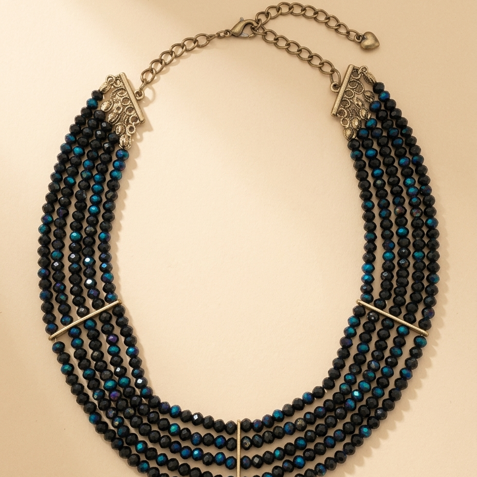 A multi-strand statement necklace featuring faceted black beads with vibrant blue iridescent accents, accented by silver-toned metal bars and chain clasp, displayed on a warm beige background.