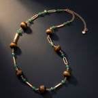 A gold-tone chain necklace featuring large brown tiger's eye beads, small green faceted beads, and elongated gold spacer bars, displayed on a dark textured background.