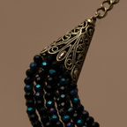 Close-up of a bohemian-style statement necklace featuring a bronze filigree cone pendant and multiple strands of faceted black beads with iridescent teal-blue reflections, set against a solid warm brown background.