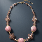 A pink beaded necklace featuring large round pink beads, faceted clear crystal beads, and gold-tone metal accents, displayed on a dark gray background.