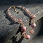 A bohemian-style beaded necklace with pink stone beads, clear faceted crystal flowers, and gold-toned metal accents, displayed on a dark textured stone surface.