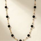 A beaded necklace featuring large glossy black round beads, gold-toned tube beads, and small gray faceted beads, arranged in a U-shape on a warm cream background.