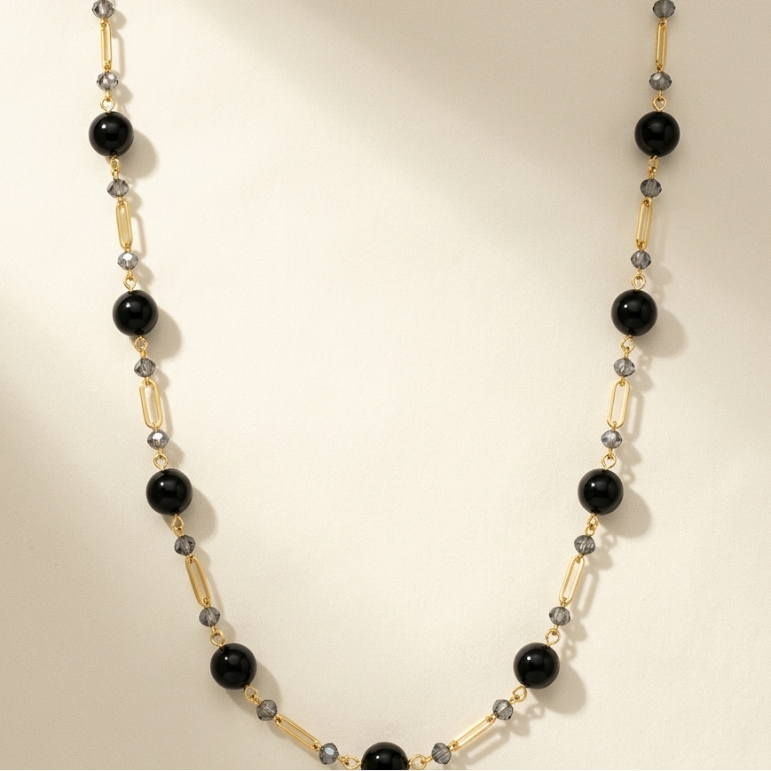 A beaded necklace featuring large glossy black round beads, gold-toned tube beads, and small gray faceted beads, arranged in a U-shape on a warm cream background.