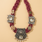 A bohemian-style statement necklace featuring a thick braided burgundy cord and ornate silver-toned metal elements with faux pearls and colorful enamel accents, displayed on a warm beige background.