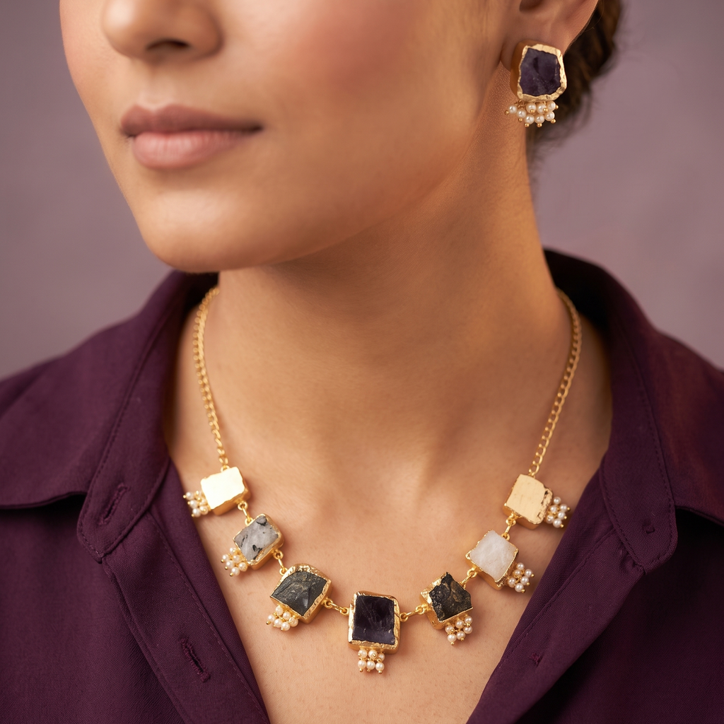 A close-up lifestyle shot of a model wearing a gold statement necklace and matching earring. The necklace features multiple rectangular stone pendants in black, white, and greenish-gray, set in gold with small pearl-like bead accents.
