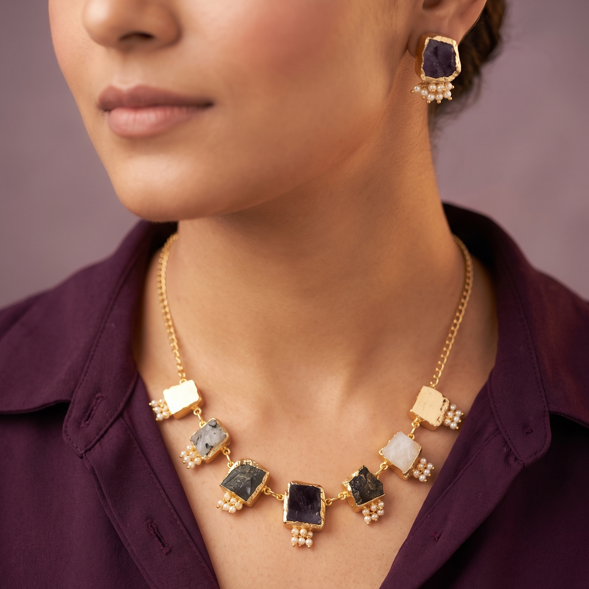 A close-up lifestyle shot of a model wearing a gold statement necklace and matching earring. The necklace features multiple rectangular stone pendants in black, white, and greenish-gray, set in gold with small pearl-like bead accents.