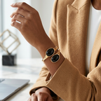A close-up lifestyle shot of a person wearing a gold-toned cuff bracelet with two large oval black stone inlays, styled with a camel blazer and white top at a marble desk.