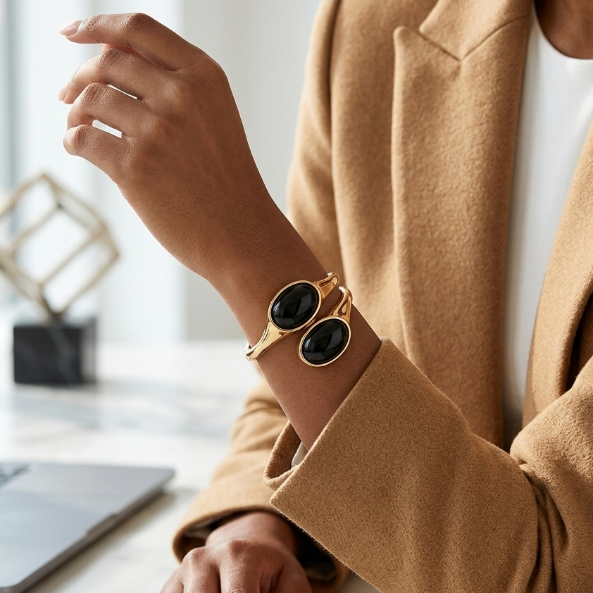 A close-up lifestyle shot of a person wearing a gold-toned cuff bracelet with two large oval black stone inlays, styled with a camel blazer and white top at a marble desk.