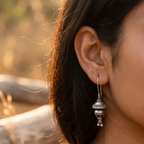 A close-up lifestyle shot of a woman's ear wearing a detailed silver-toned traditional Indian pendant earring with a small hanging bell, set against a warm, blurred outdoor background.