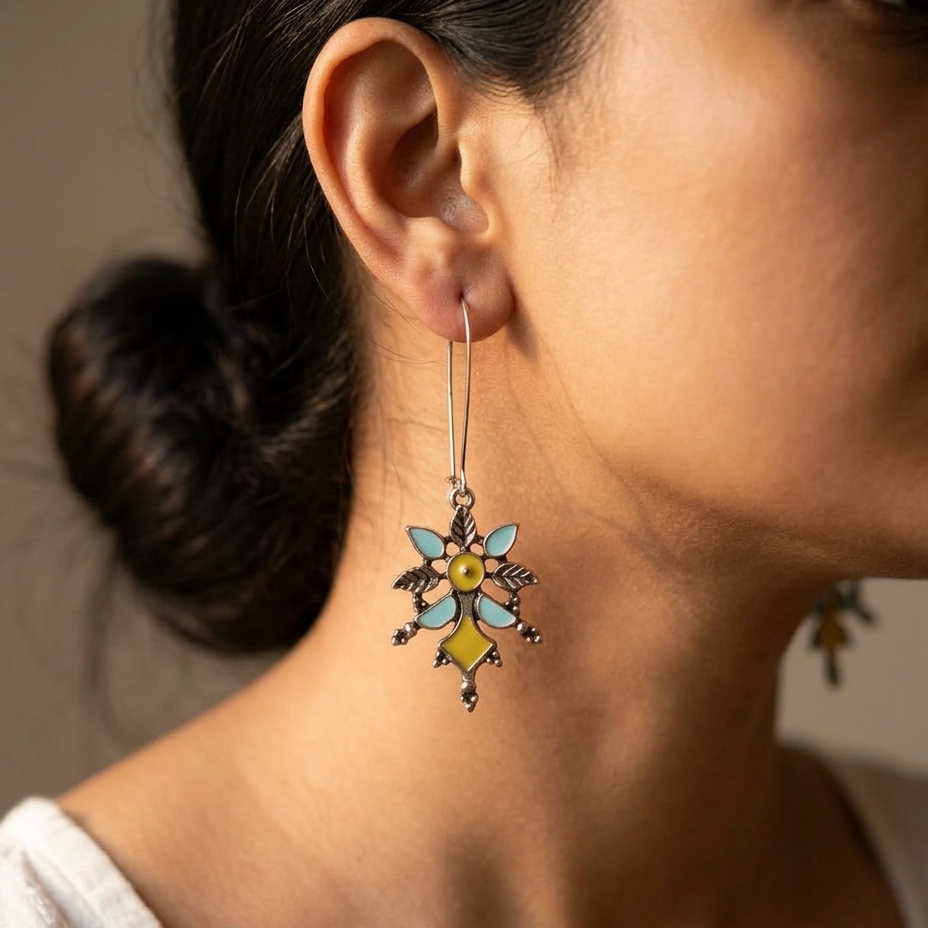 A close-up lifestyle shot of a woman wearing a dangling earring. The earring features a silver-toned metal frame with a colorful enamel design resembling a stylized flower or star, with light blue, yellow, and orange details.
