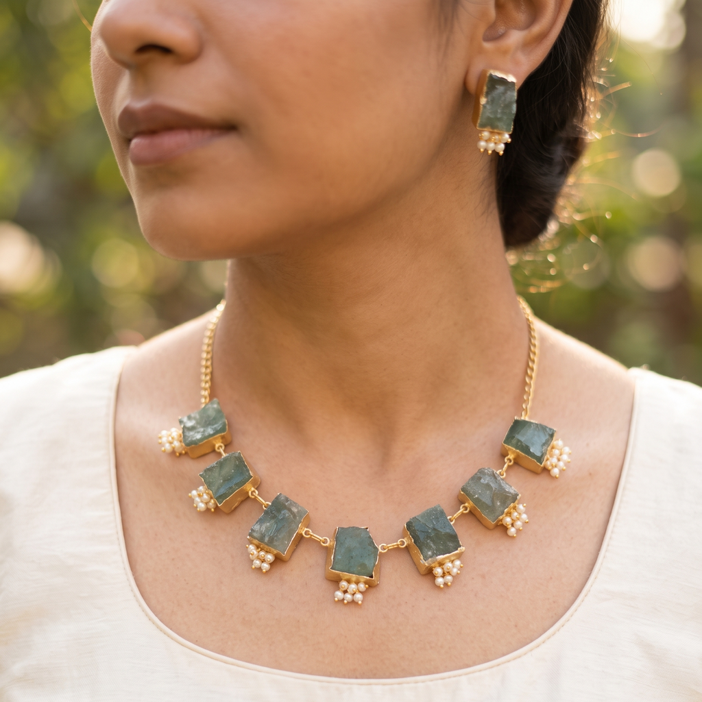 A close-up lifestyle shot of a woman wearing a gold-plated statement necklace and matching drop earring. The necklace features rectangular green stone inlays set in gold, accented with small gold and pearl bead clusters.