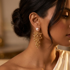 A close-up lifestyle shot of a woman wearing a gold dangle earring with a single white pearl at the top, styled with her hair in an elegant low bun and wearing a cream satin one-shoulder dress.