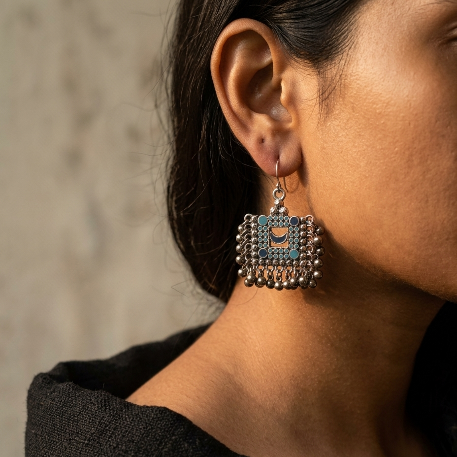 A close-up lifestyle shot of a woman wearing a square silver-toned dangle earring with intricate blue and gold enamel details and dangling silver beads.