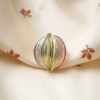 A close-up of a decorative ring shaped like a leaf, featuring a central gold-toned leaf flanked by silver and rose gold leaf segments, resting on draped cream satin fabric with embroidered leaf motifs.