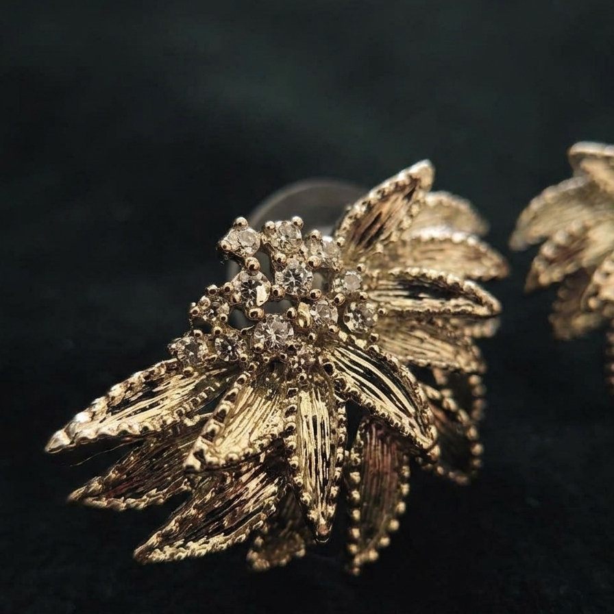 A close-up of a gold-toned statement earring with textured leaf-like petals and clear rhinestone accents, photographed on a dark black background. Part of a second matching earring is visible at the right edge.