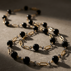 A close-up of a gold and black beaded necklace with alternating matte black round beads and polished gold oval links, draped on a textured gray surface with dramatic shadows.