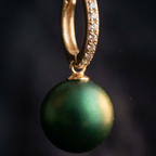 A close-up of a gold huggie hoop earring adorned with small clear pavé stones and featuring a dangling oval green pearl, set against a dark, soft-focus background.