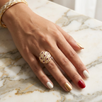 A close-up of a hand resting on a white and gray marble surface, wearing a chunky gold ring with a textured, diamond-like pattern and a matching gold bracelet.