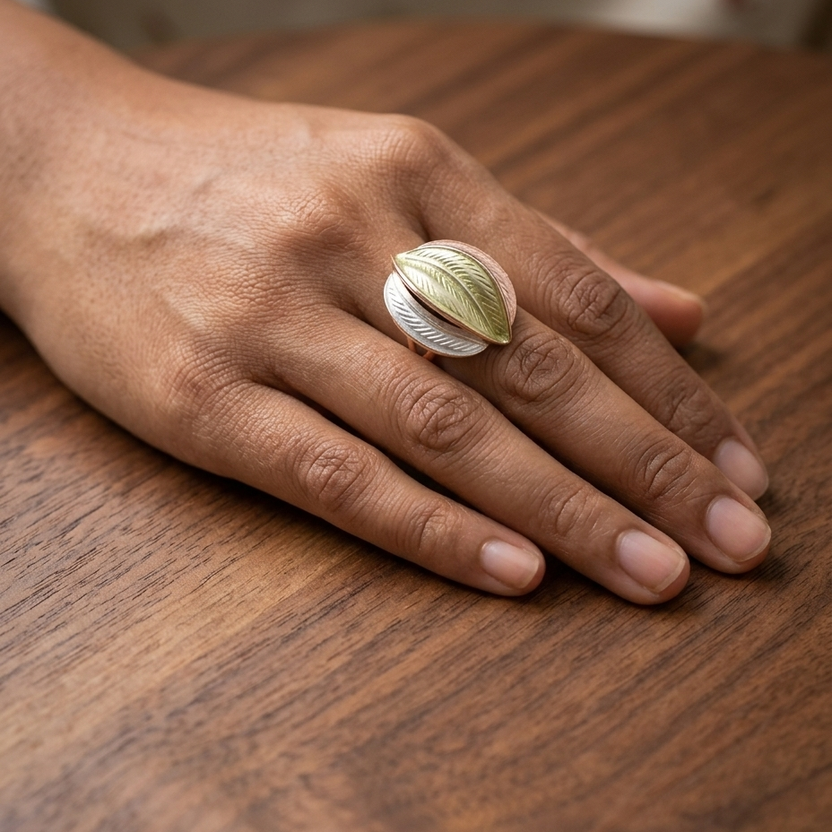 A close-up of a hand resting on a wooden table, wearing a sculptural ring shaped like a textured green leaf layered over a white striped cylindrical base.