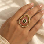A close-up of a hand wearing a large teardrop-shaped ring with a pink stone, white pearl-like border, and gold detailing, set against a soft cream fabric background.
