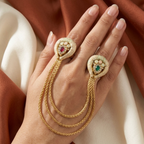 A close-up of a hand wearing a pair of ornate gold ringlets with decorative heart-shaped tops, one accented with a pink gemstone and the other with a green gemstone, set against draped rust and cream fabric.