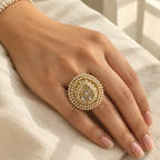 A close-up of a hand wearing an ornate gold ring decorated with rows of white pearls and small green and white stones, resting on a cream-colored fabric surface.
