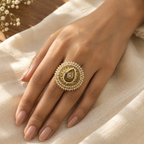 A close-up of a hand wearing an ornate gold ring decorated with small white pearls and a central teardrop-shaped stone, resting on draped cream fabric.
