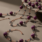 A close-up of a jewelry strand with large purple beads, rose gold chain links, and small dark gray faceted beads, arranged on a warm taupe surface with a matching curved pedestal.