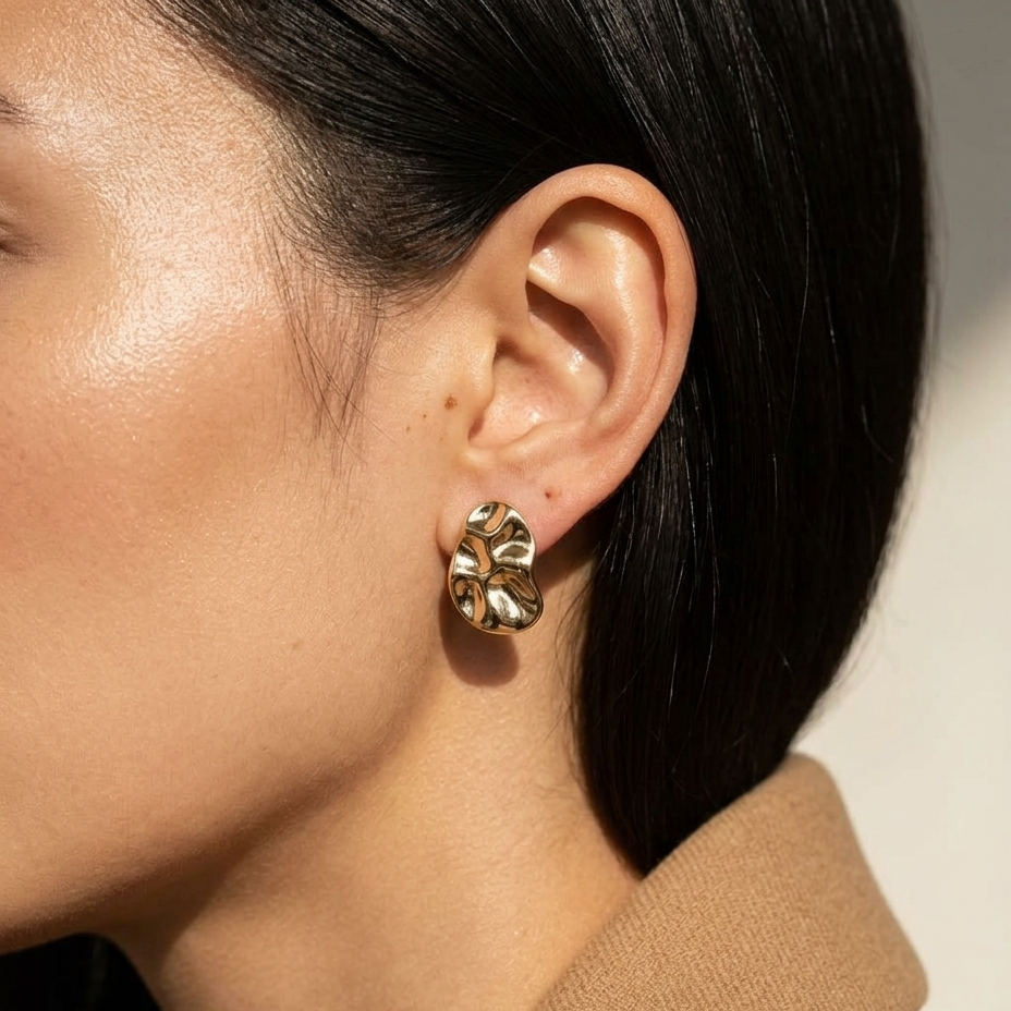 A close-up of a model's ear wearing a textured, organic-shaped gold stud earring, with her dark hair and tan jacket collar visible against a neutral light background.
