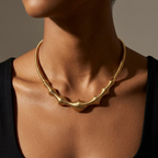 A close-up of a model's neck and chest wearing a sculptural, wavy gold necklace over a black top, set against a neutral warm gray background.