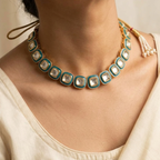 A close-up of a model wearing a gold-toned traditional Indian-style necklace with square turquoise and white stone inlays, along with a matching earring and cream-colored top.