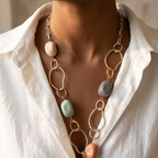 A close-up of a model wearing a gold chain necklace with abstract oval links and pastel-colored stone-like beads in beige, sage green, peach, and gray, styled with a white linen shirt.