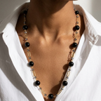 A close-up of a model wearing a long necklace with black beads, gold paperclip links, and clear crystal accents over a white collared shirt.