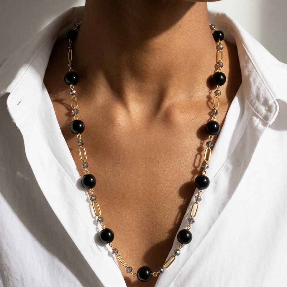 A close-up of a model wearing a long necklace with black beads, gold paperclip links, and clear crystal accents over a white collared shirt.