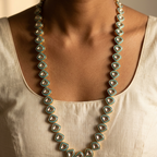 A close-up of a model wearing a long statement necklace made of repeating turquoise and gold geometric beads over a cream-colored linen top.