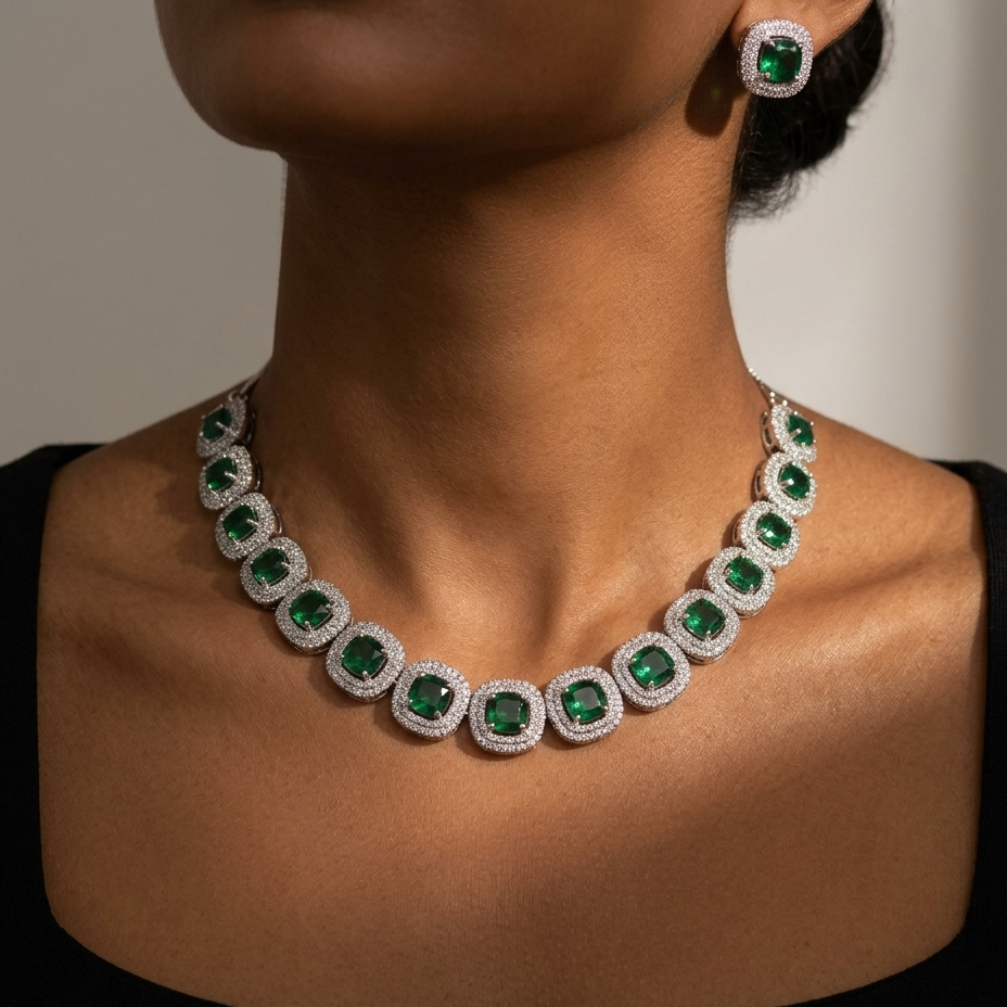 A close-up of a model wearing a silver-toned necklace and matching stud earring, both featuring square-cut green emeralds surrounded by pavé-set clear stones.