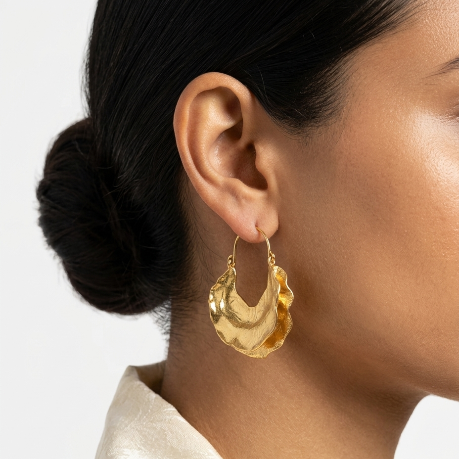 A close-up of a model wearing a textured, organic-shaped gold hoop earring. The model has dark hair styled in a bun and is wearing a cream satin shirt against a clean white background.