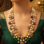 A close-up of a model wearing a traditional Indian multi-strand statement necklace. The necklace features white pearls, gold-plated beads, yellow and green stone accents, and red stone details, paired with a dark green embroidered blouse.