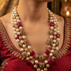 A close-up of a model wearing an ornate traditional Indian gold-plated necklace with white faux pearls, magenta-pink cube gemstones, and small green and red accent stones, paired with a red embroidered blouse.