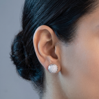 A close-up of a person's ear wearing a textured silver stud earring, with dark hair pulled back and a neutral gray background.