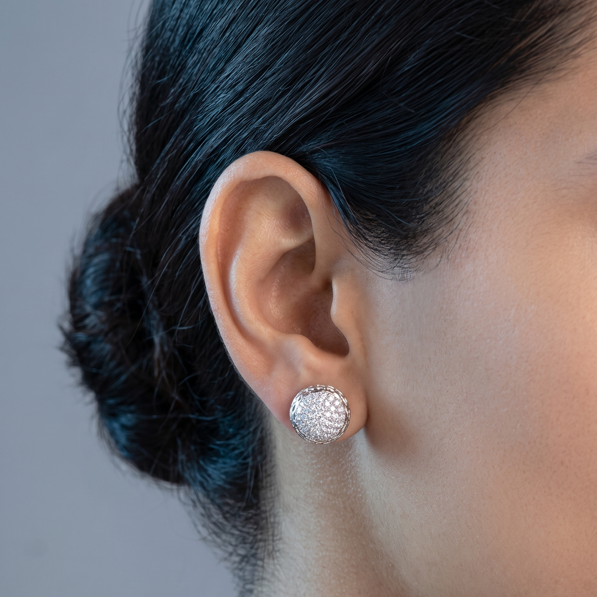 A close-up of a person's ear wearing a textured silver stud earring, with dark hair pulled back and a neutral gray background.