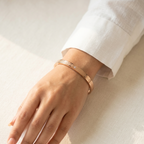 A close-up of a person's hand and wrist wearing a slim rose gold bangle bracelet with a textured woven pattern and a small clear stone, resting on a light beige linen surface.