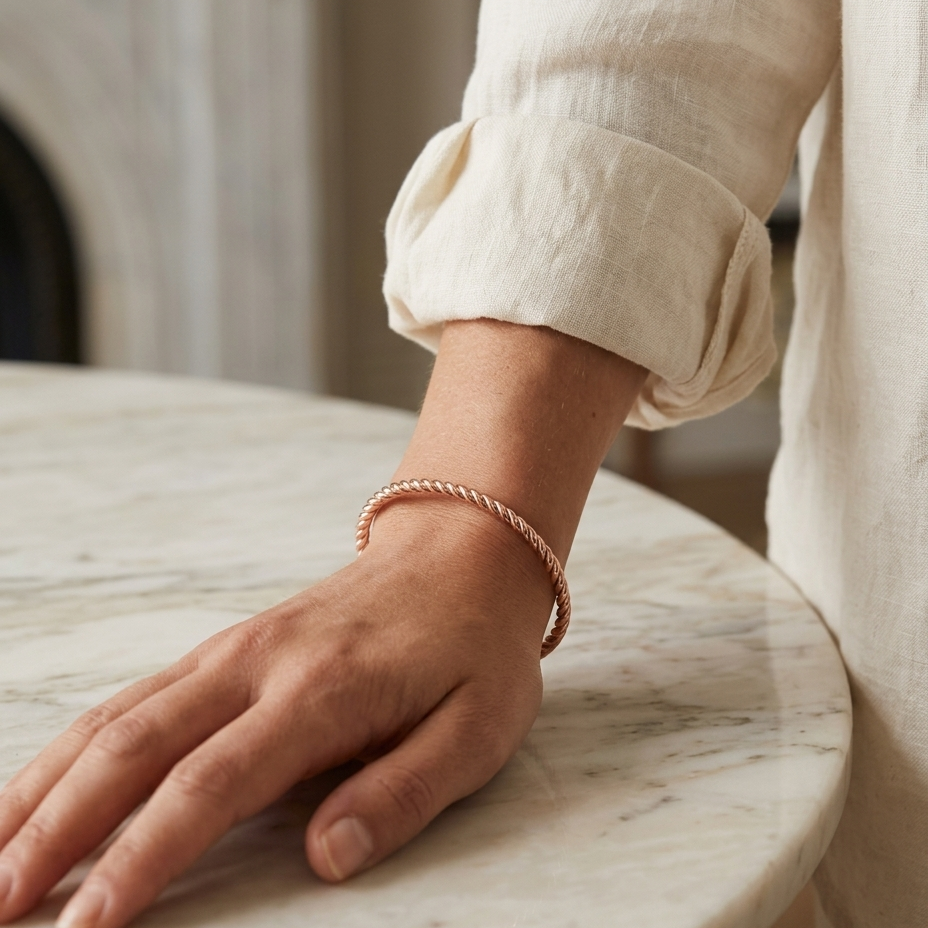 A close-up of a person's hand resting on a white marble table while wearing a thin, twisted rose gold bangle bracelet.