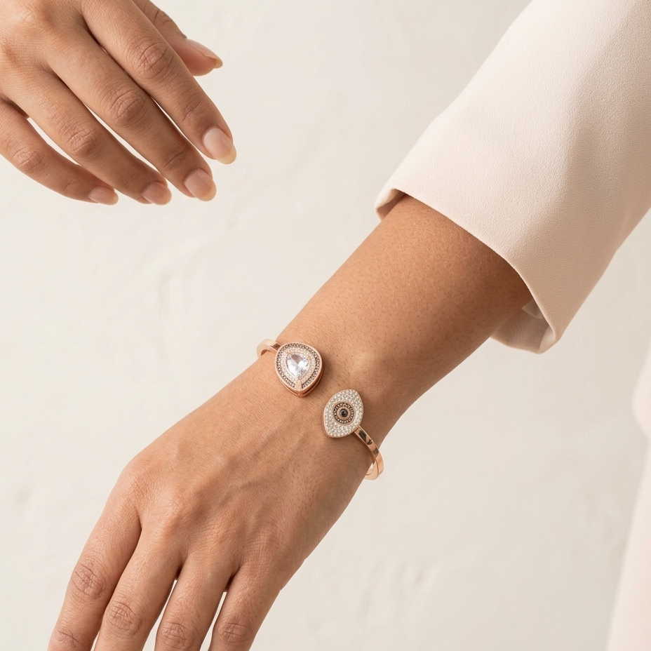 A close-up of a person's wrist and hand wearing a delicate rose gold cuff bracelet with two circular charms, one heart-shaped and one eye-shaped, both adorned with clear stones. The person is wearing a light beige sleeve, and the background is a clean, textured off-white wall.