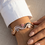 A close-up of a person's wrist wearing a decorative wavy cuff bracelet. The bracelet has a rose gold base with black enamel sections, white floral motifs, and small colorful circular accents.