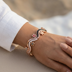 A close-up of a person's wrist wearing a rose gold cuff bracelet with a black enamel wave, small colorful evil eye motifs, and clear crystal details, set against a soft beige background.