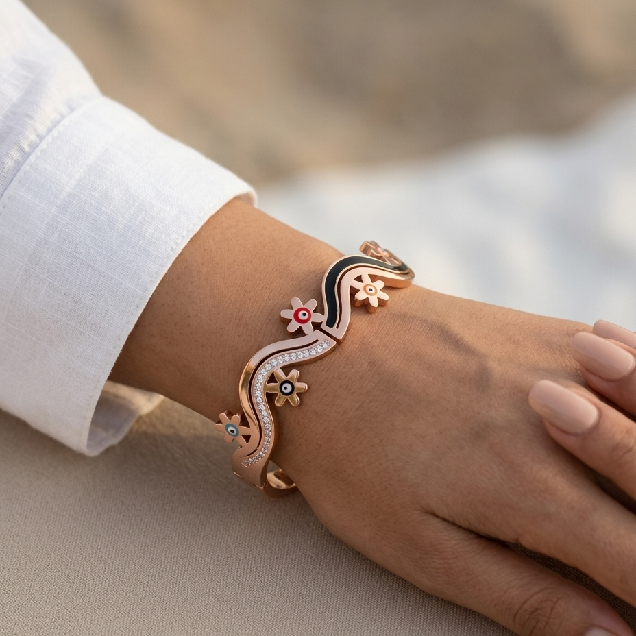 A close-up of a person's wrist wearing a rose gold cuff bracelet with a black enamel wave, small colorful evil eye motifs, and clear crystal details, set against a soft beige background.