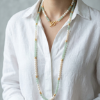 A close-up of a person wearing a long double-strand beaded necklace with white pearls, light green jade-like beads, and small gold spacer beads, layered over a gold pendant necklace with a toggle clasp, paired with a white linen shirt.
