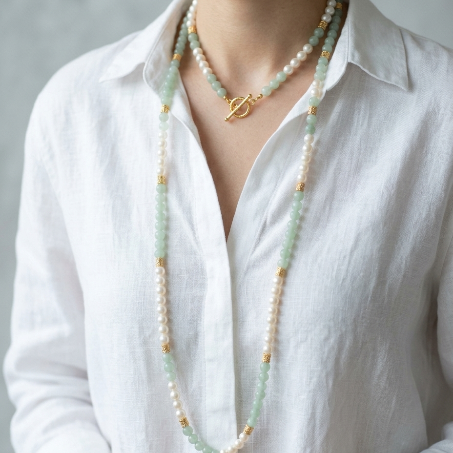 A close-up of a person wearing a long double-strand beaded necklace with white pearls, light green jade-like beads, and small gold spacer beads, layered over a gold pendant necklace with a toggle clasp, paired with a white linen shirt.